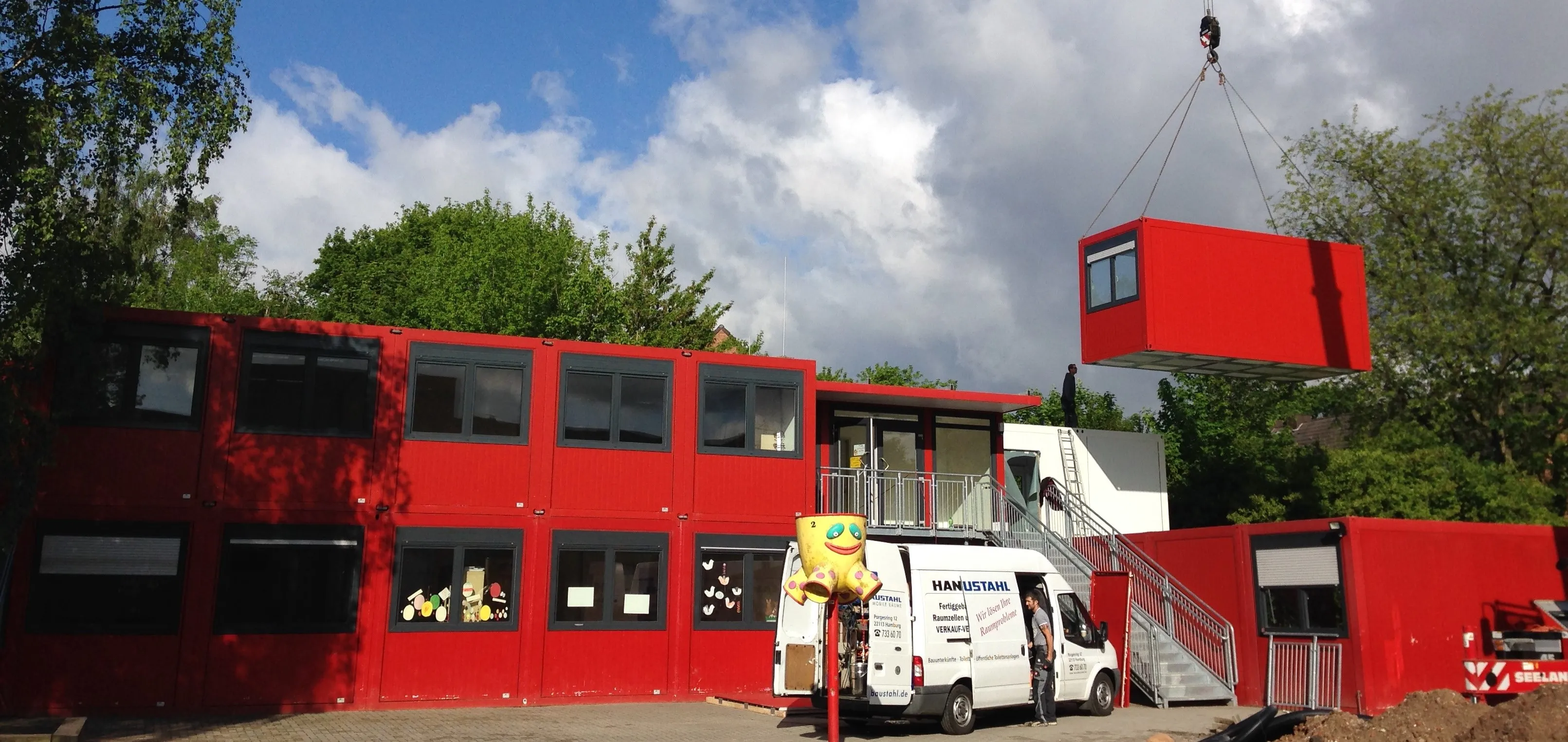 Rote modulare Schulgebäude aus Containern werden mit einem Kran montiert, davor steht ein weißer Transporter und eine bunte Dekofigur.