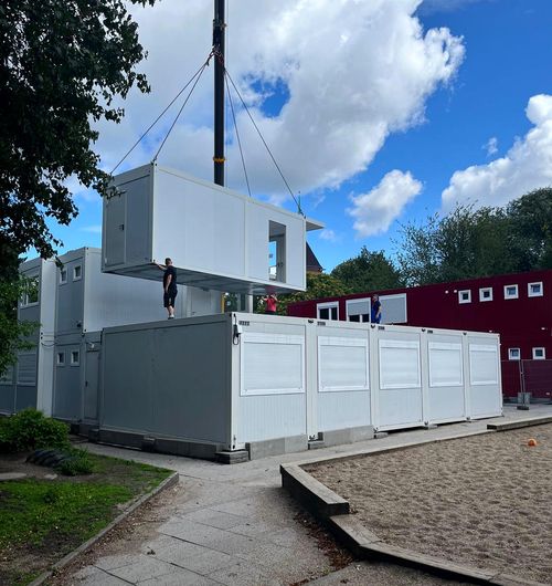 Ein Kran hebt einen weißen modularen Container über gestapelte Container auf einem Schulhof bei klarem Himmel.