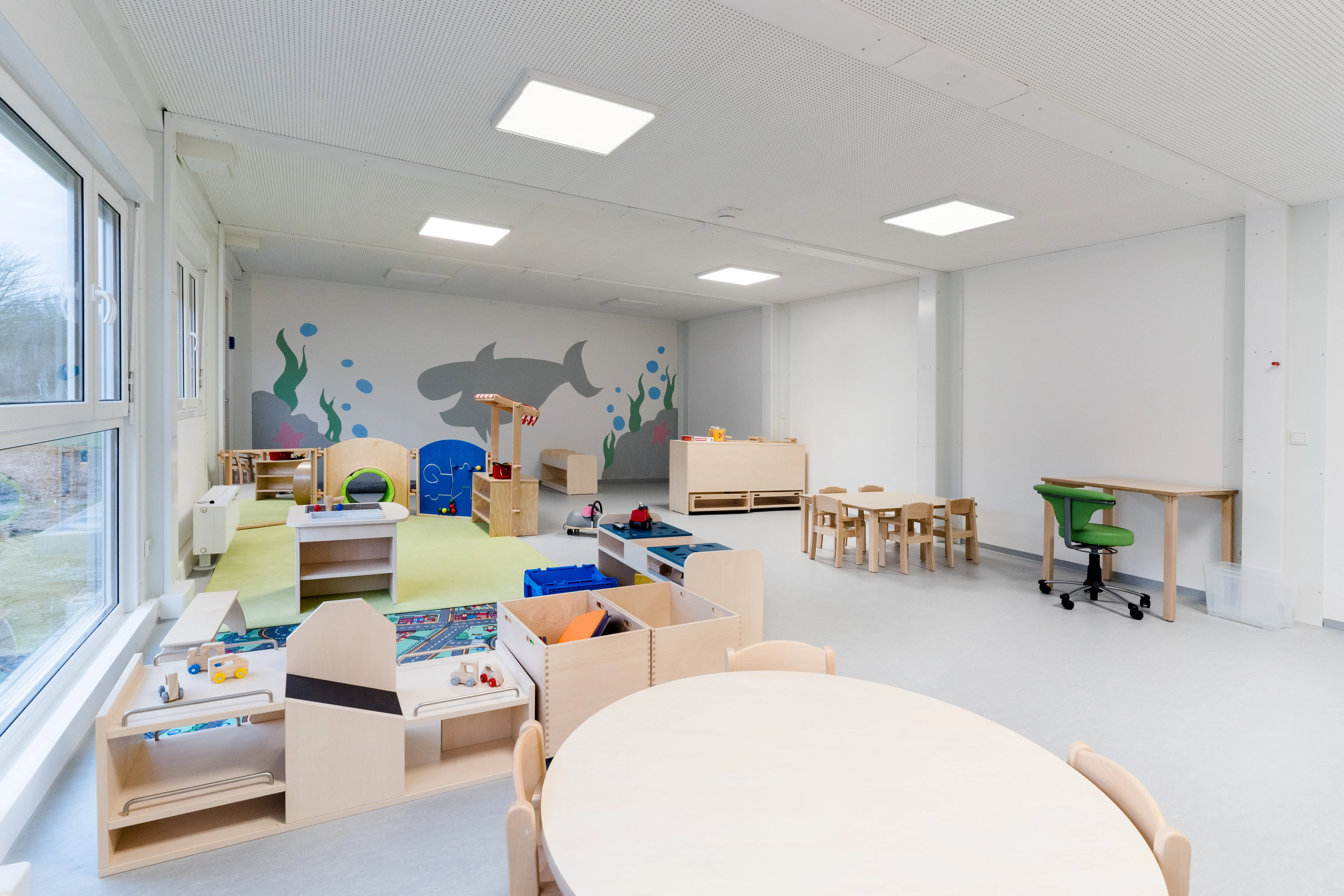 Helle und moderne Kindertagesstätte mit bunten Spielbereichen, Holzmöbeln und Meerestier-Wandmalerei.