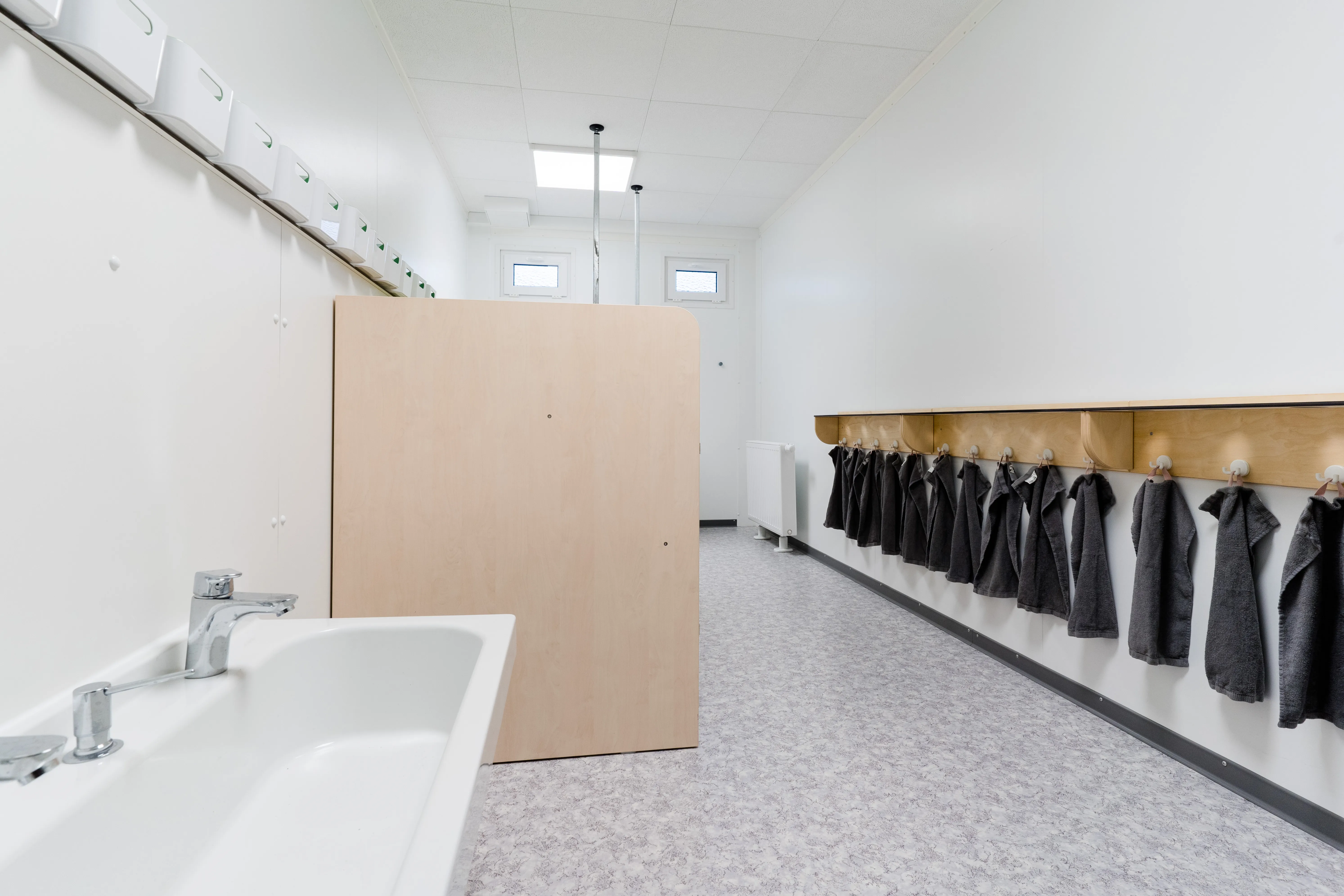 Hell beleuchteter Waschraum mit zwei Waschbecken, Holzwandtrennung und aufgereihten kleinen dunkelgrauen Handtüchern an Haken entlang der Wand.