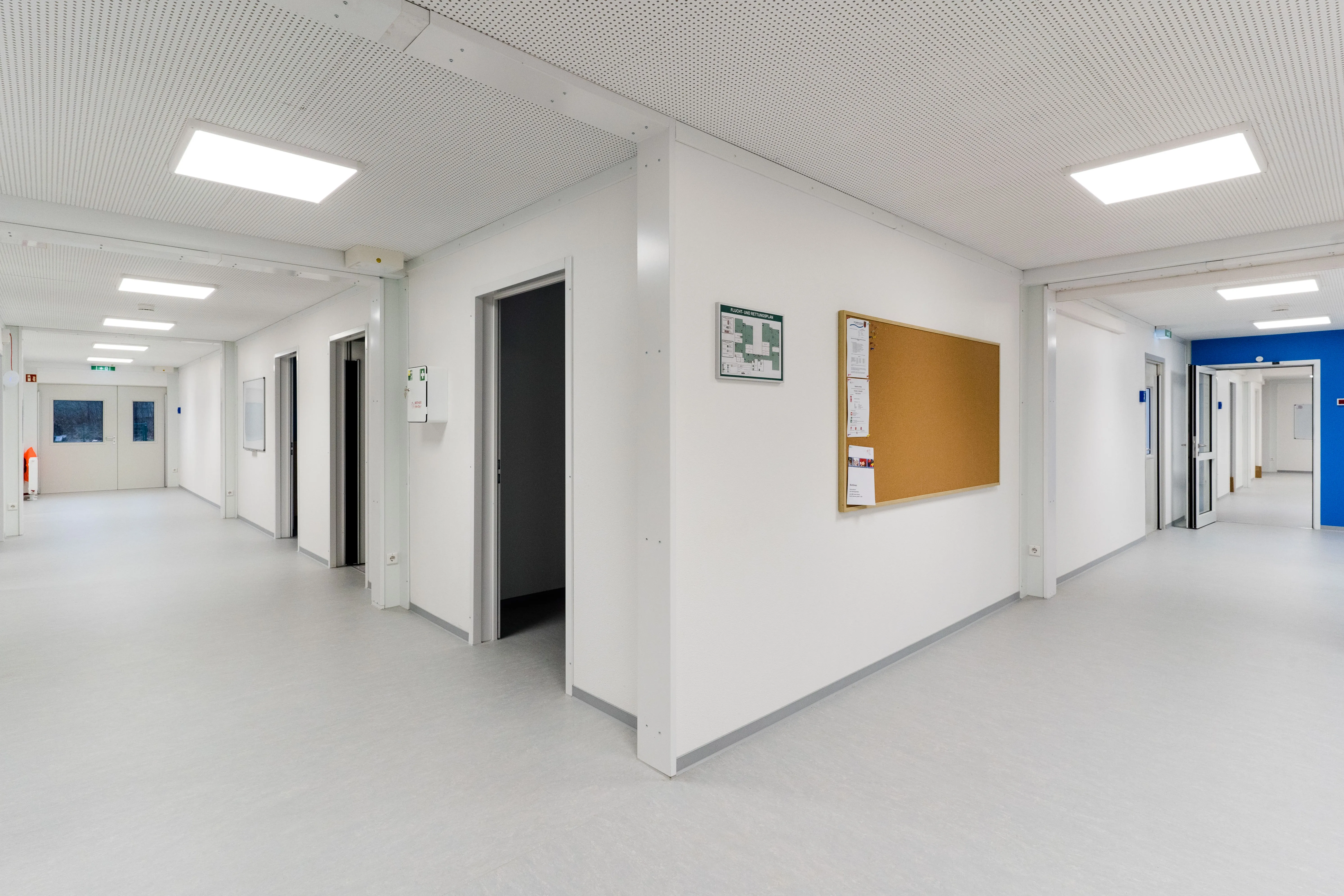 Moderne, helle Flure mit weißen Wänden und mehreren offenen Türen, beleuchtet von rechteckigen Deckenleuchten.