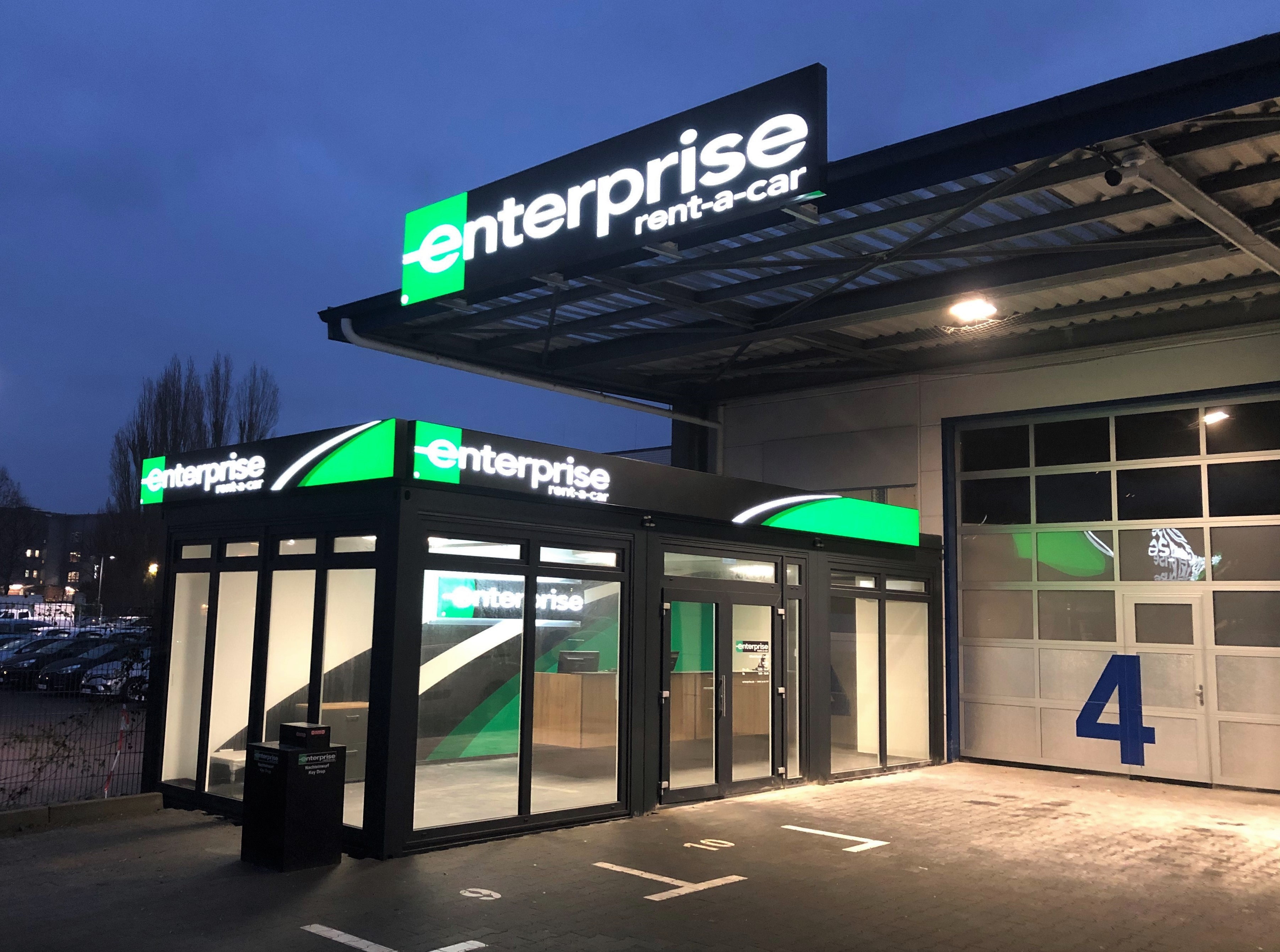 Enterprise Rent-a-Car Verleihstation bei Dämmerung mit beleuchtetem grünen und weißen Logo auf einem modernen Glasgebäude.