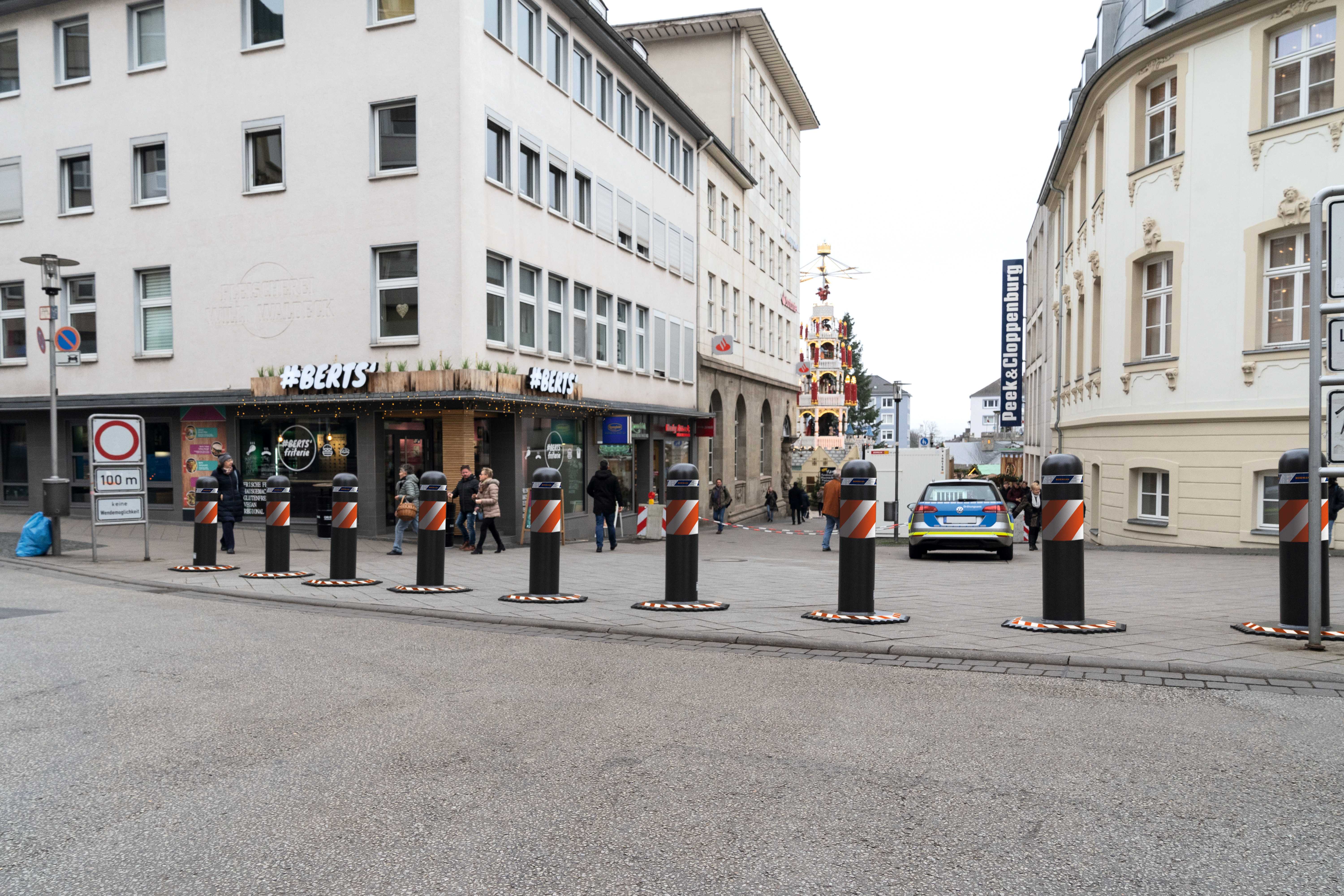 Straßenszene mit schwarzen Absperrpfosten, mehreren Fußgängern, einem Polizeiauto und Gebäuden mit Geschäften, darunter #BERTS und Peek & Cloppenburg.