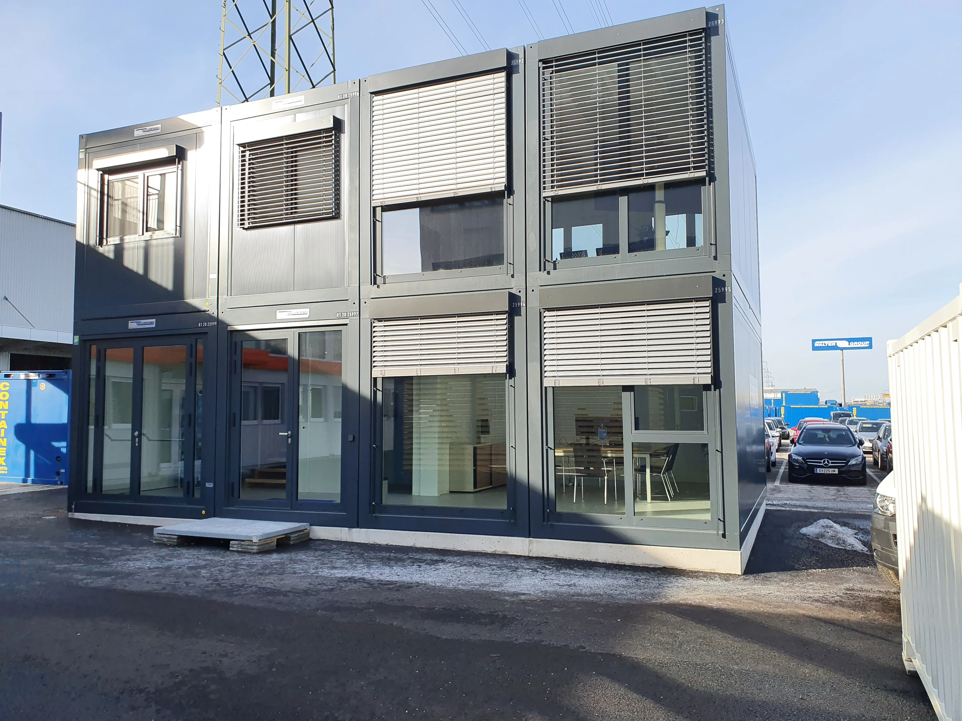 Modulare Bürocontainer mit großen Fenstern und Jalousien, stehen auf einem Parkplatz bei klarem Himmel.