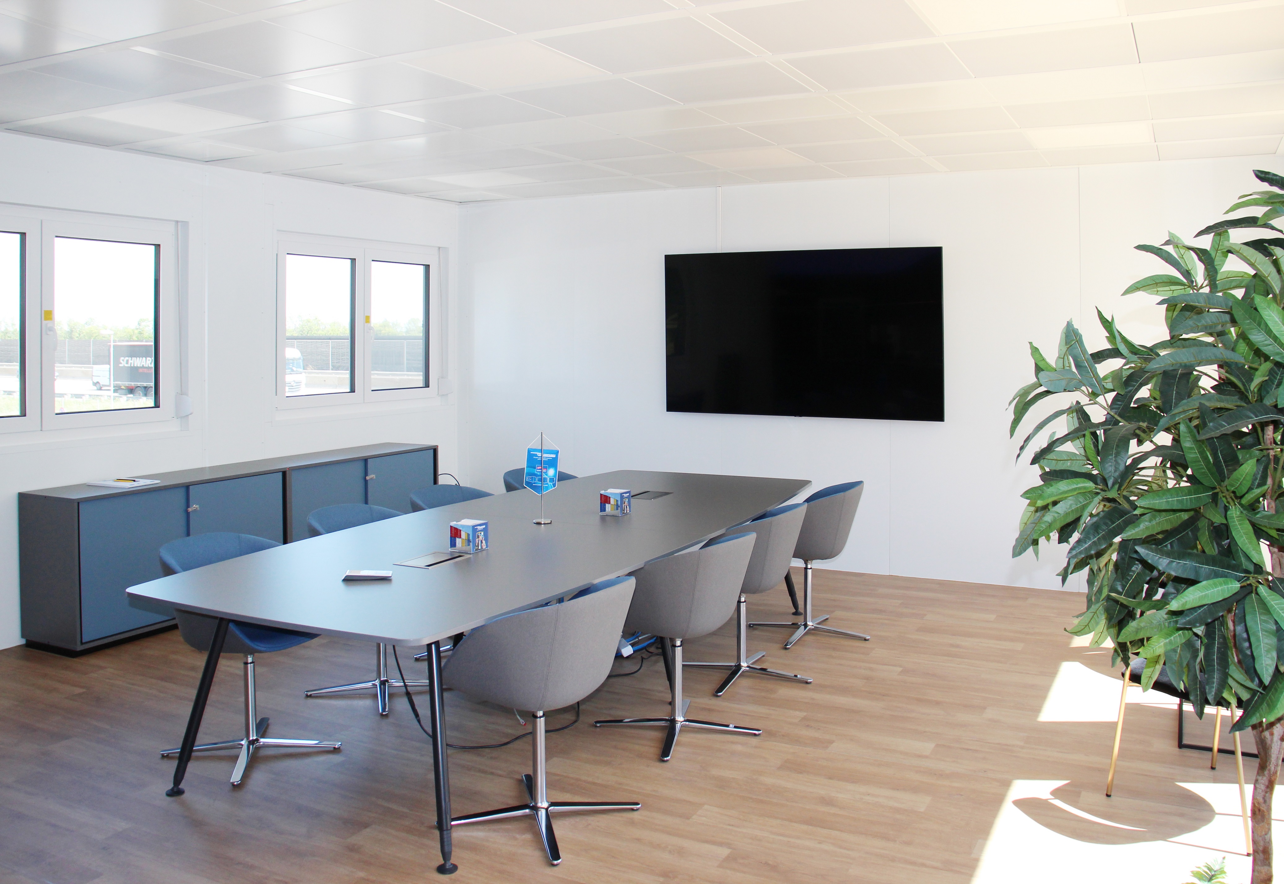 Moderner Besprechungsraum mit langem Tisch, mehreren Stühlen, großem Bildschirm an der Wand und Pflanzen in der Ecke.