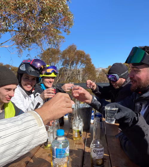Group of people wearing ski helmets and goggles sitting at a wooden table outdoors, raising glasses in a toast under a clear blue sky.