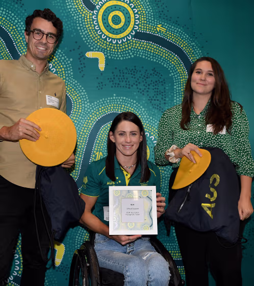 Three people smiling in front of a patterned green background, one woman in a wheelchair holding a certificate, flanked by a man and another woman holding yellow hats and navy bags.