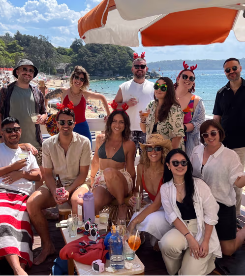 Group of friends wearing festive red antler headbands gathered on a beachside patio with drinks, under an orange and white umbrella.
