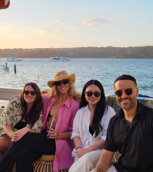 Four friends wearing sunglasses sitting and smiling near a waterfront with yachts in the background.