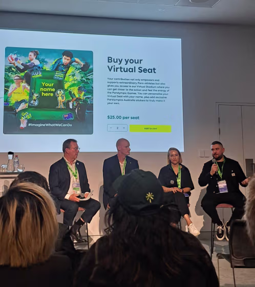 Four people seated on a panel in front of an audience with a presentation slide titled 'Buy your Virtual Seat' showing a fundraising campaign featuring Paralympic athletes.