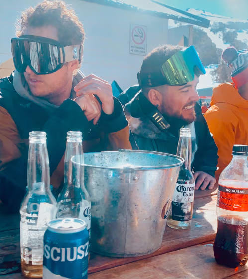 Two men wearing ski goggles sitting at a wooden table outdoors with empty beer bottles, a metal bucket, and a soda bottle under bright sunlight.