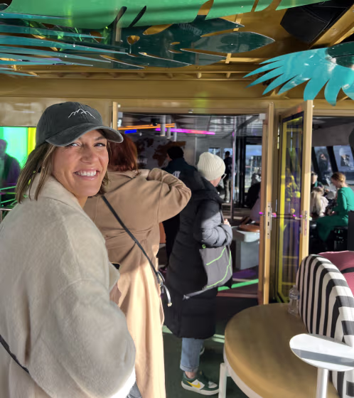 Smiling woman wearing a baseball cap and beige coat inside a room with people, decorative green and gold leaves hanging from the ceiling.