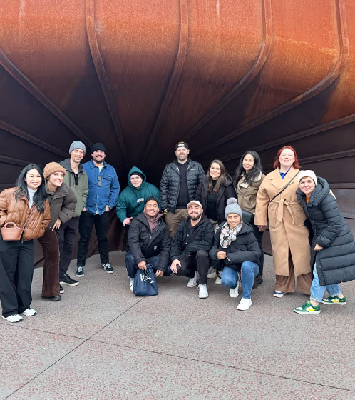 Group of thirteen people wearing winter clothes posing and smiling outdoors under a large rust-colored metal structure.