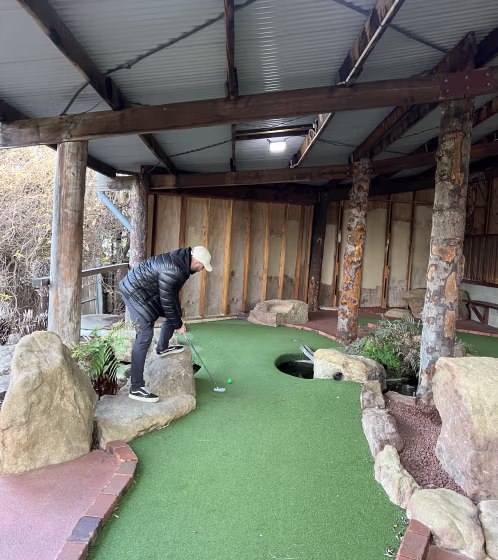 Person in a black jacket and white cap playing mini-golf on an indoor course with artificial turf and stone edges.