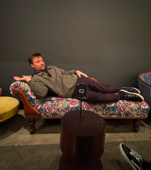 Man lying sideways on a floral patterned chaise lounge indoors with a dark wall background.