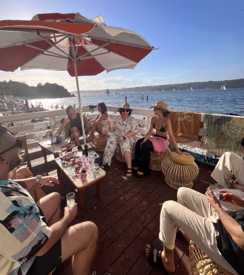 Group of people sitting and relaxing under a large umbrella on a wooden deck overlooking a beach and ocean with sailboats in the distance.