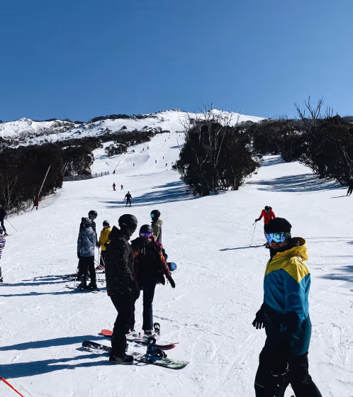 Group of snowboarders and skiers on a snowy slope with a clear blue sky and tree-lined mountains in the background.