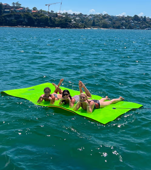 Three children lying and relaxing on a large bright green floating mat in a body of water with a shoreline in the background.