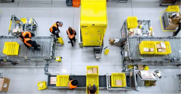 Workers wearing orange safety vests sorting and moving yellow bins on conveyors in a warehouse.