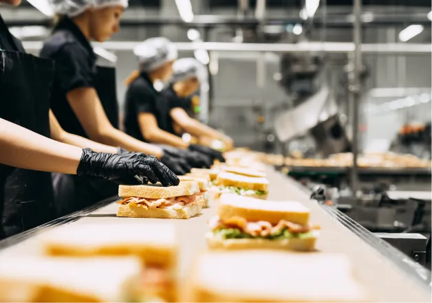 Workers wearing hairnets and black gloves assembling sandwiches on an industrial production line.