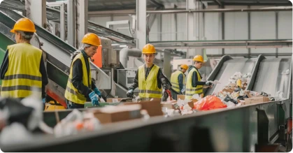 Workers wearing yellow safety vests and helmets sorting recyclable materials on a conveyor belt in a recycling facility.