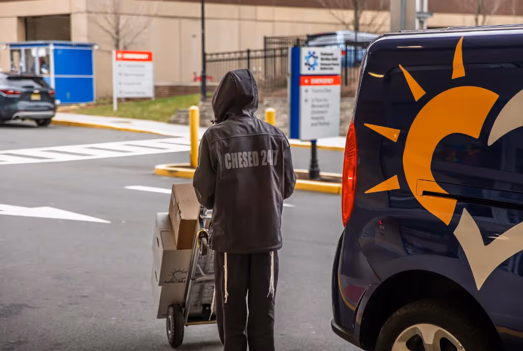 Chesed 24/7 staff member loading medical supplies onto a cart beside a shuttle vehicle outside a New York City hospital