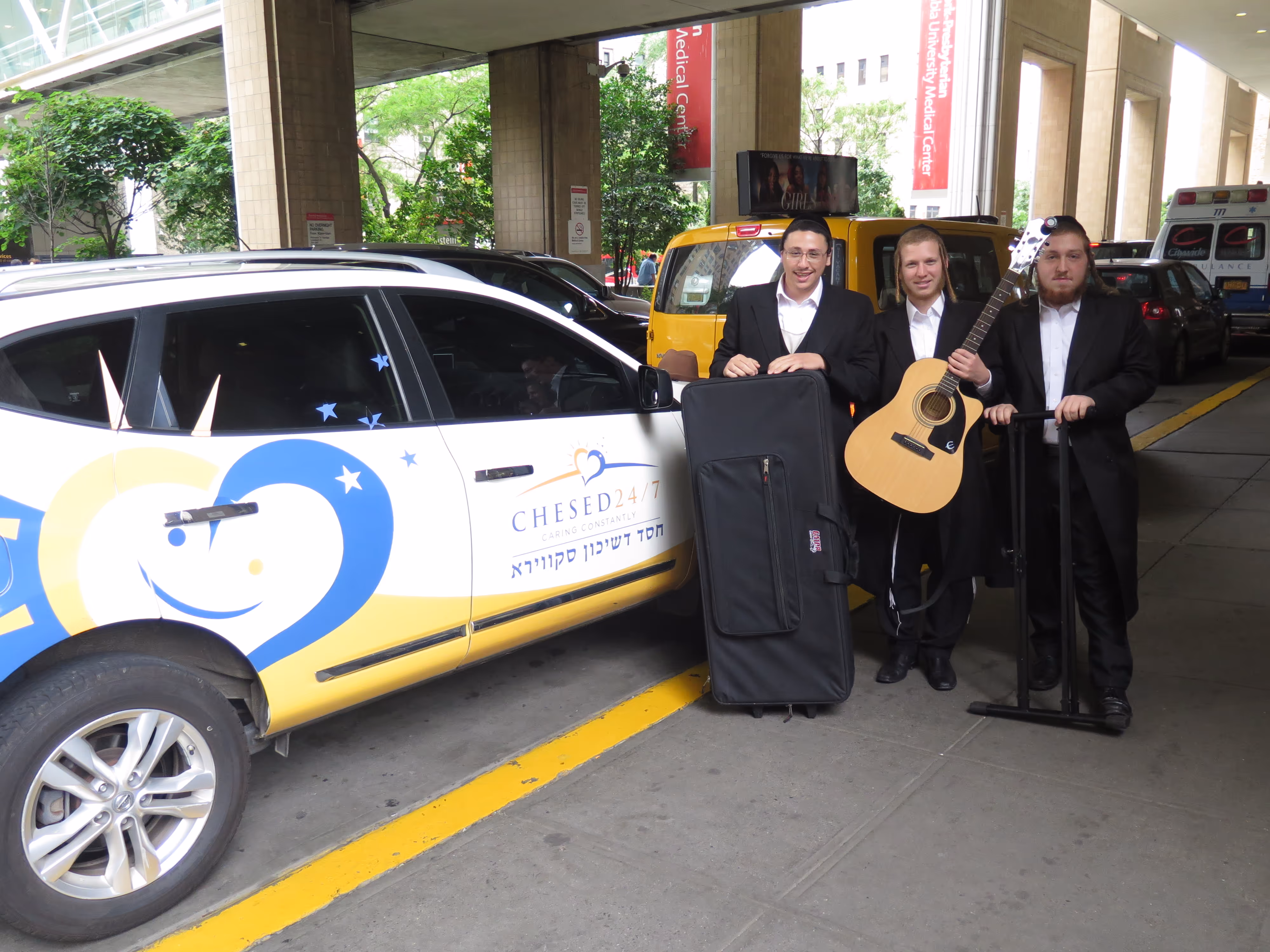 Smile 24/7 volunteers with musical instruments standing beside a Chesed 24/7 vehicle outside a hospital, preparing to bring music and emotional support to patients