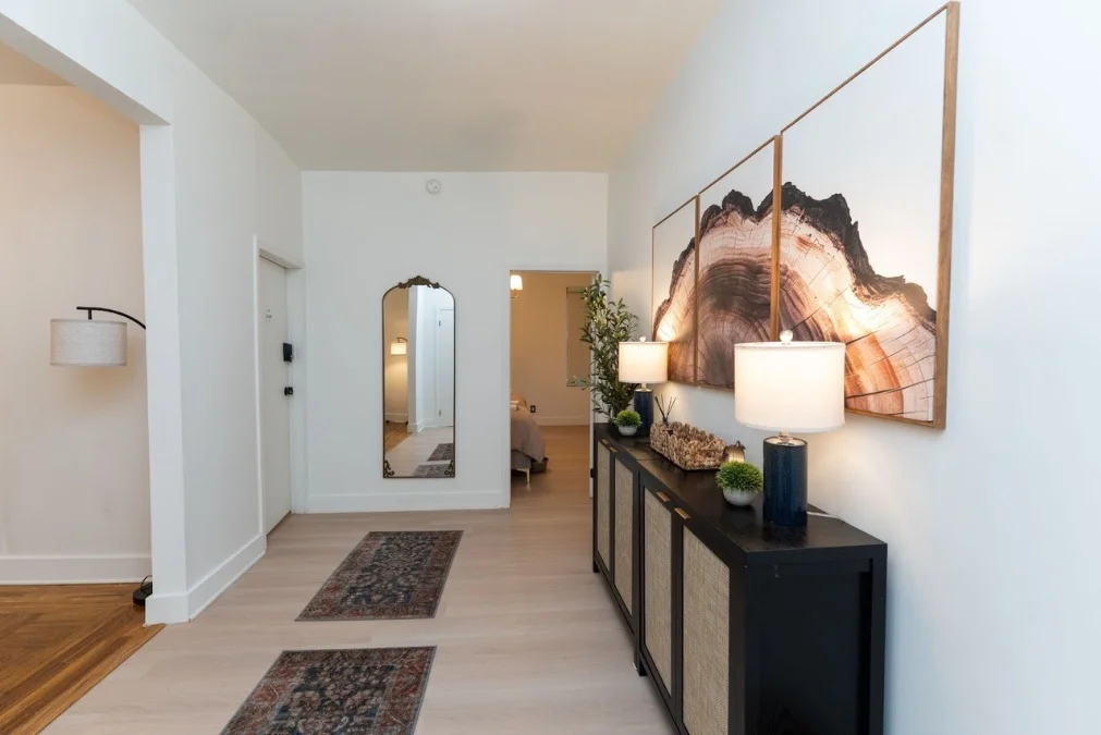 Furnished hallway inside the Chesed 24/7 apartment near Columbia Presbyterian Hospital featuring warm lighting, decorative console table, wall art, and bedroom entrance