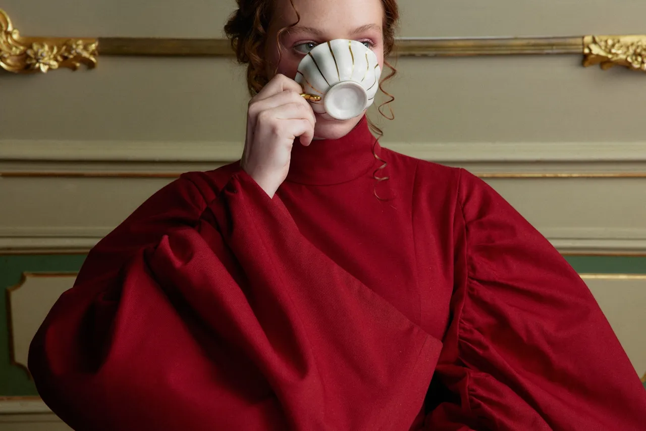 Woman in a voluminous red dress sipping from a white teacup with gold lines in an ornate room.