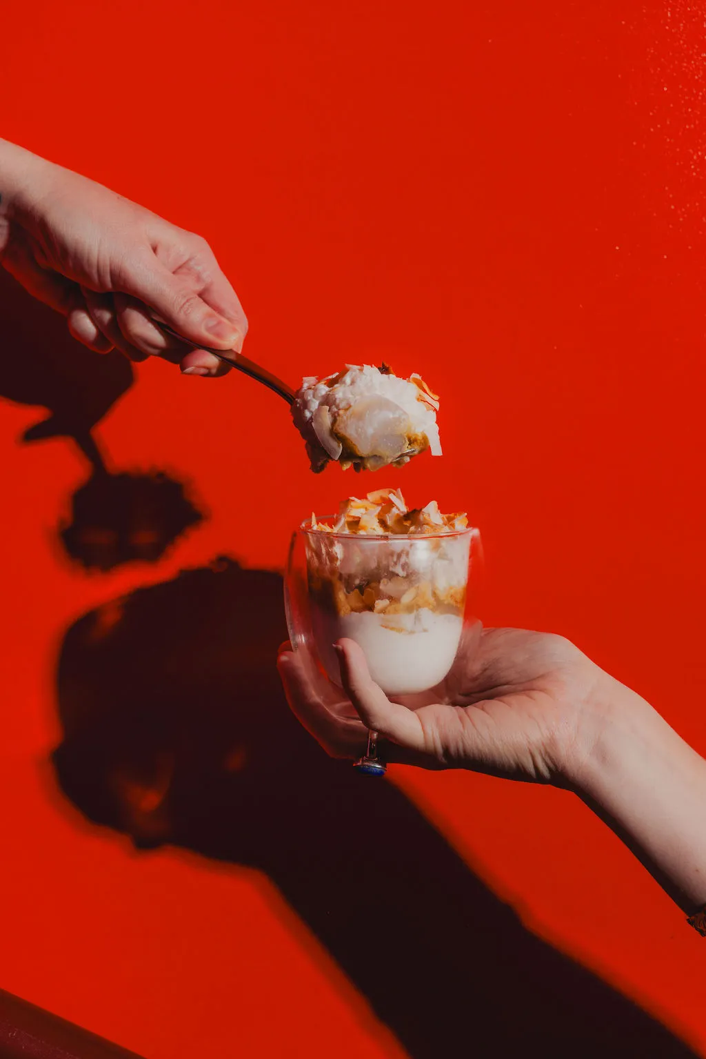 Hands holding a glass cup filled with layered dessert and a spoon lifting a portion against a red background.