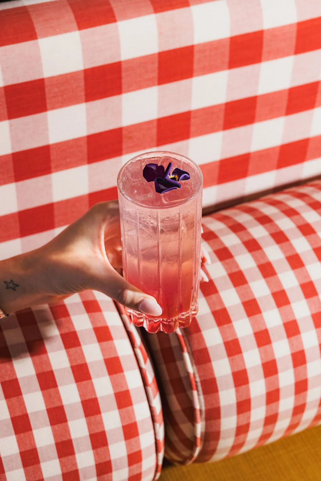 Hand holding a pink iced drink garnished with a small purple flower against a red and white checkered background.