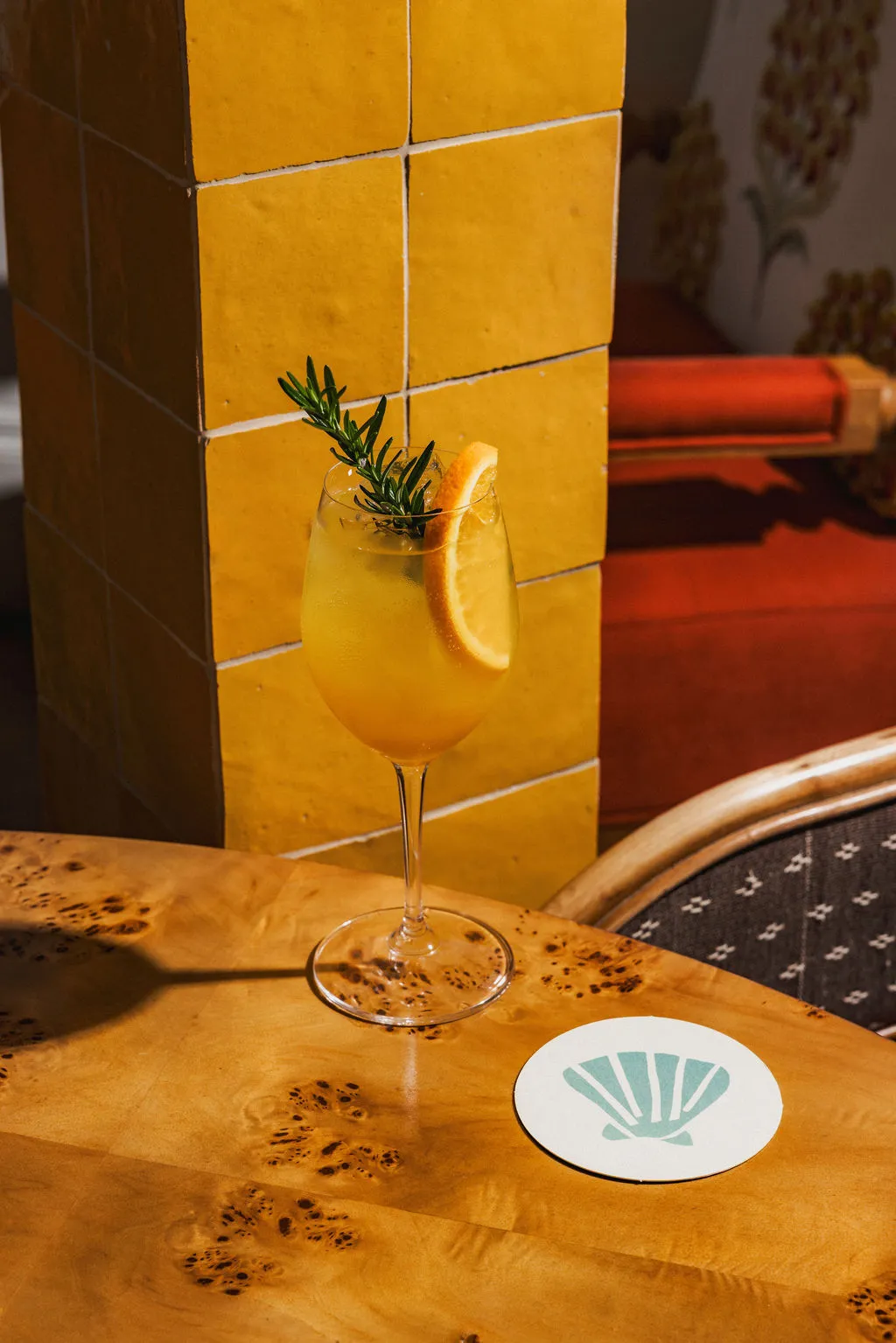 Yellow cocktail garnished with a rosemary sprig and orange slice on a wooden table next to a yellow tiled column.