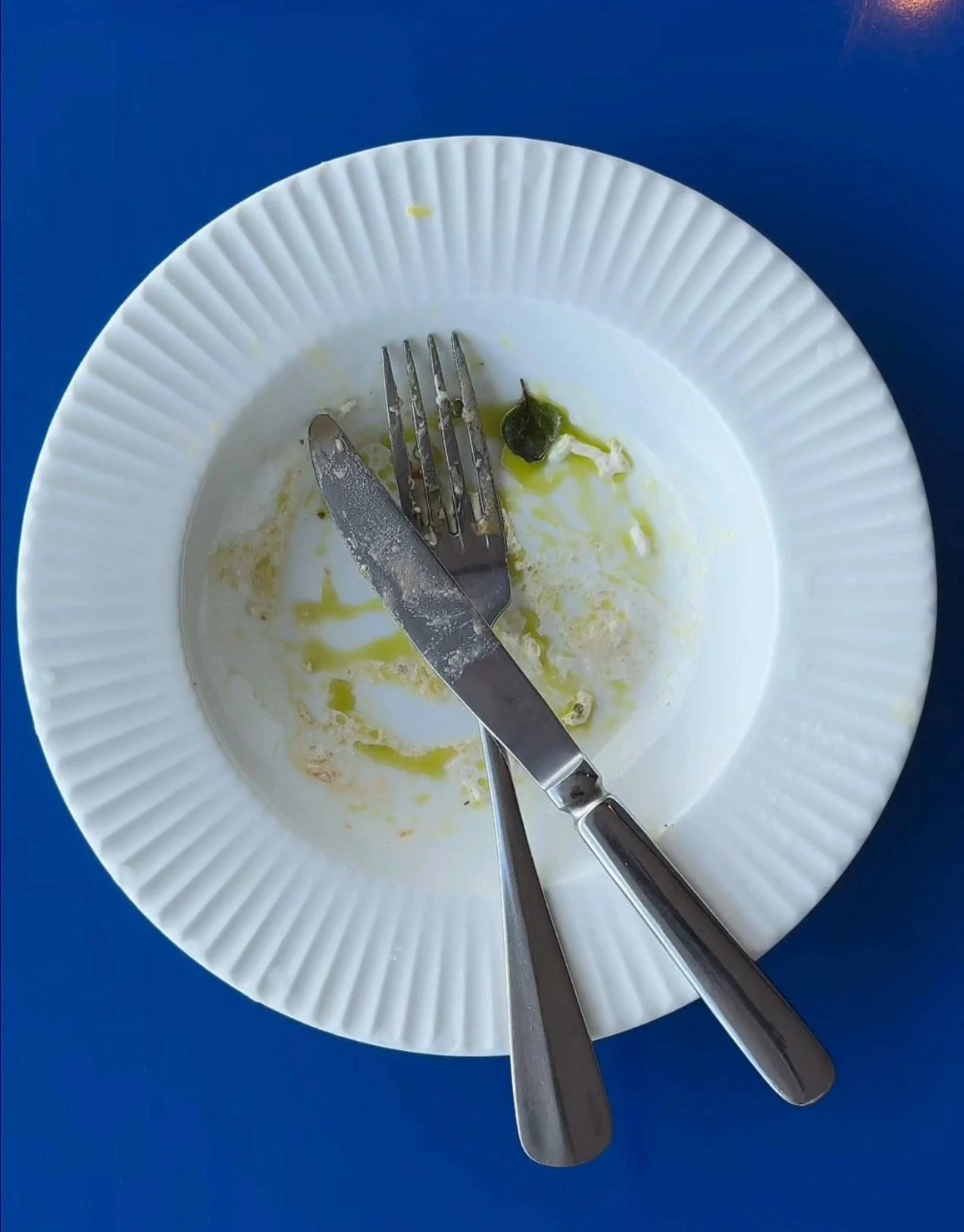 Empty white plate with knife and fork crossed on it and leftover sauce and a small green leaf on a blue surface.