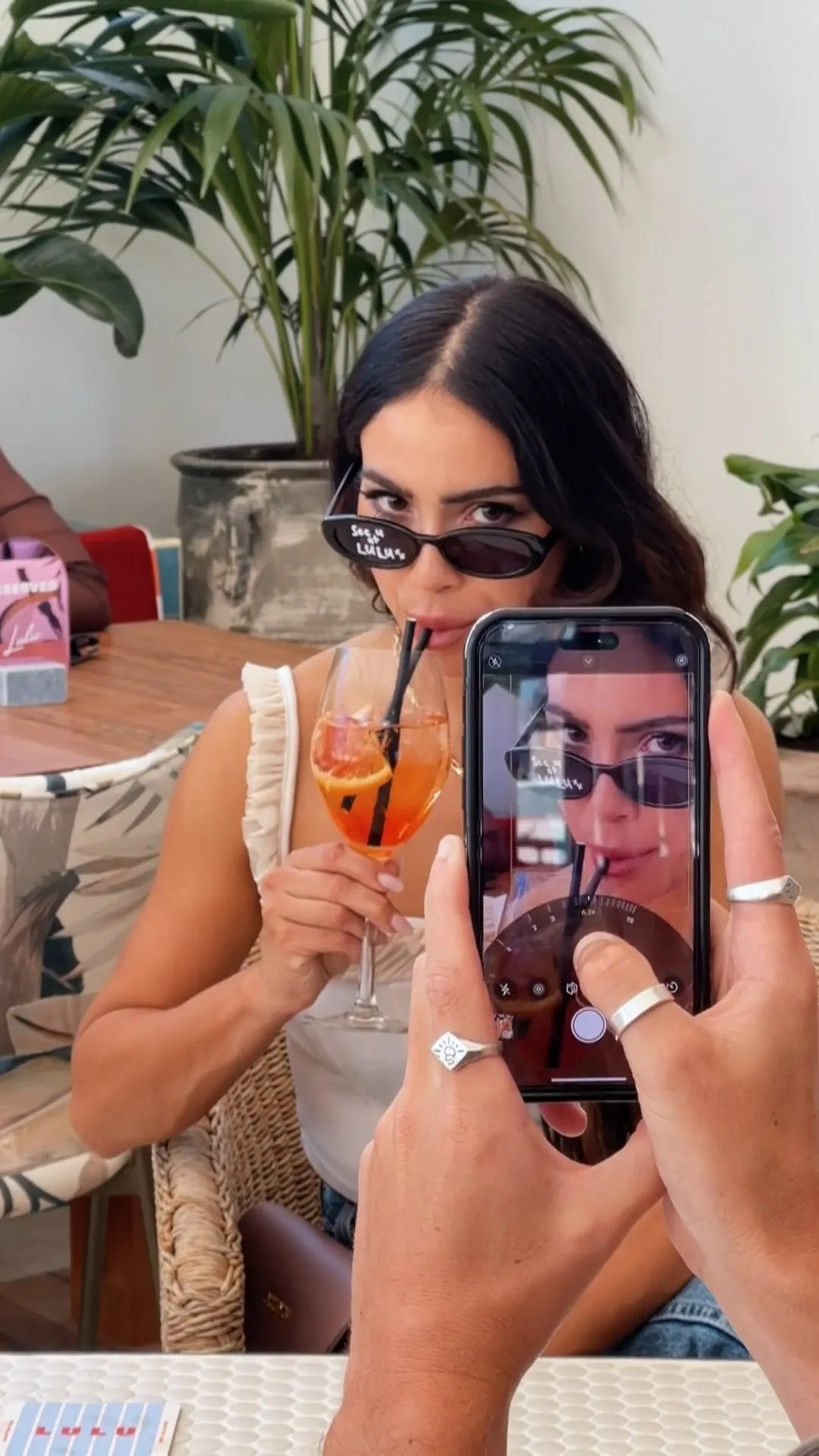 Woman with dark hair wearing sunglasses drinking an orange cocktail through two straws, being photographed on a smartphone.