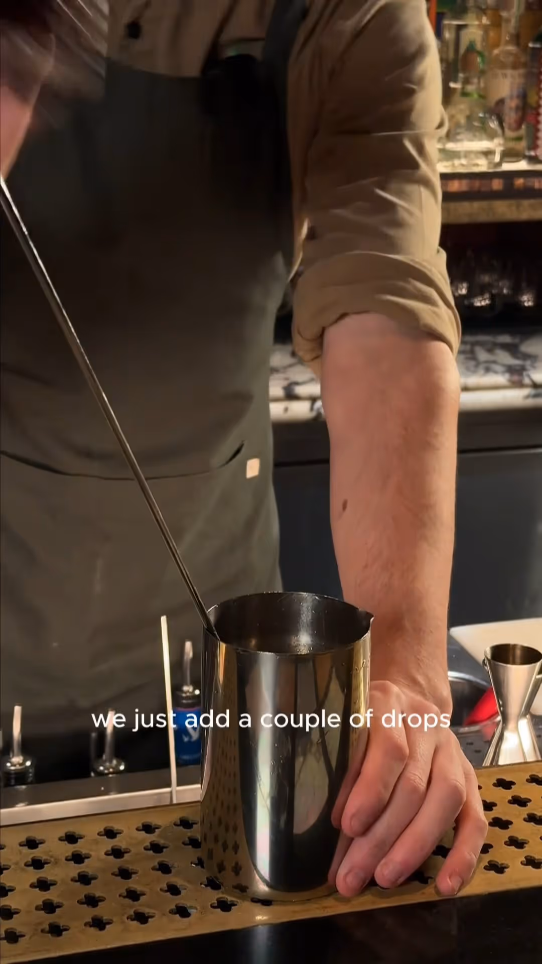 Bartender wearing apron stirring a drink in a metal shaker on a bar counter.