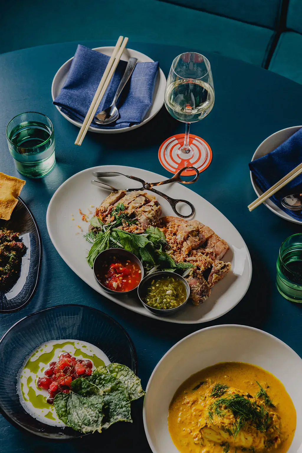 Table with a meal consisting of sliced roasted meat with herbs and two sauces, a bowl of creamy yellow curry garnished with herbs, a dish with creamy white sauce, red diced topping and green leaf crisps, glass of white wine, and glass of water with chopsticks and blue napkins nearby.