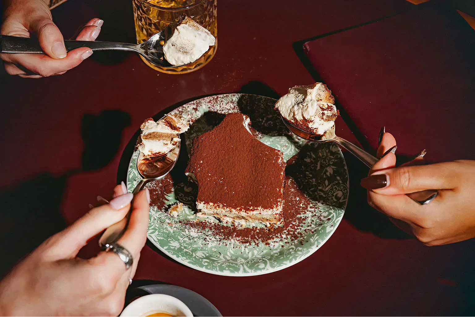 Three hands holding spoons with tiramisu dessert around a partially eaten tiramisu slice on a floral plate on a dark red table.