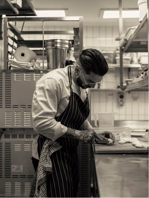 Tattooed chef wearing glasses and apron slicing food on a cutting board in a professional kitchen.