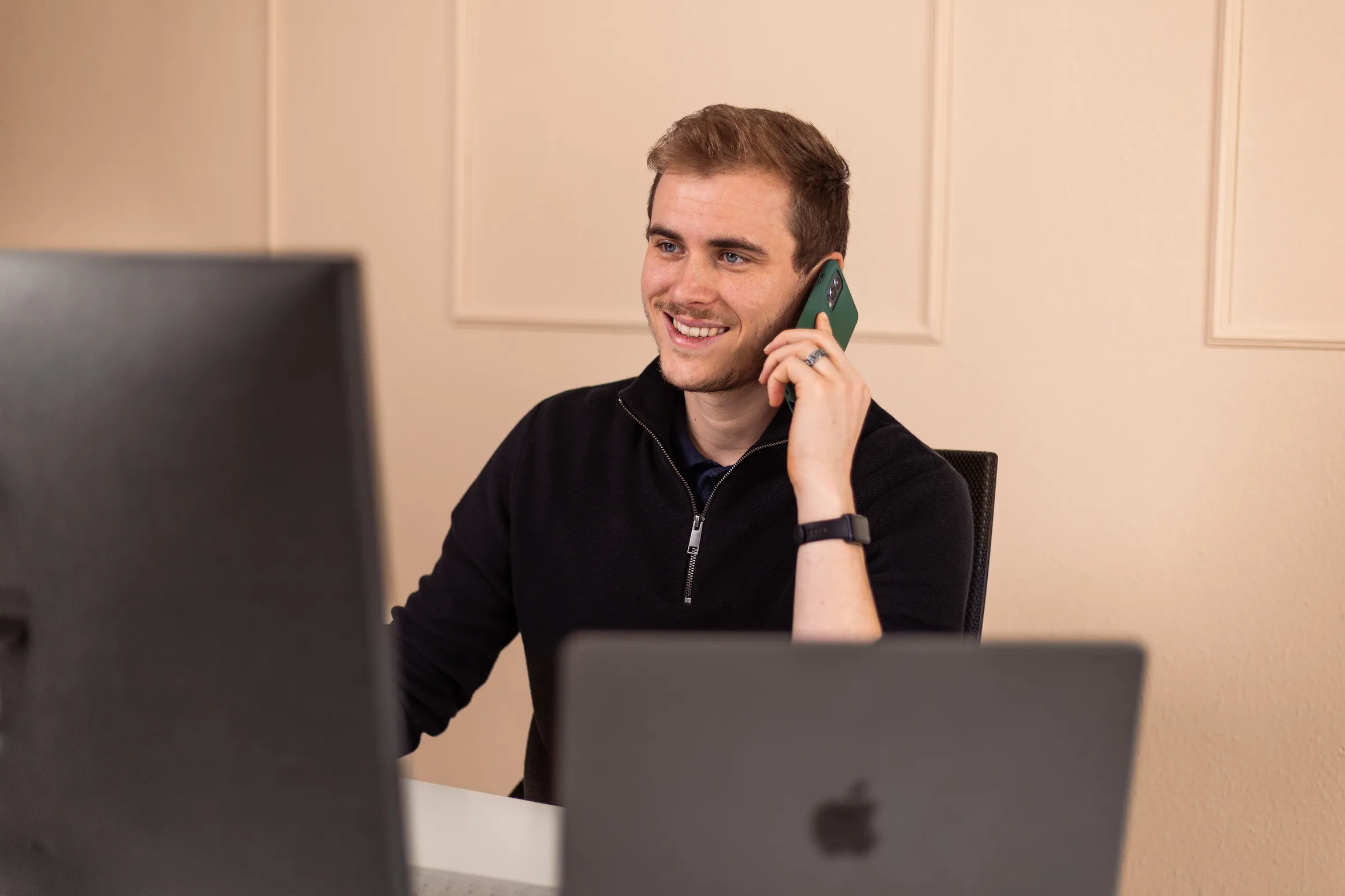 Junger Mann mit schwarzem Pullover spricht am Telefon, sitzt an Schreibtisch mit zwei Computermonitoren.
