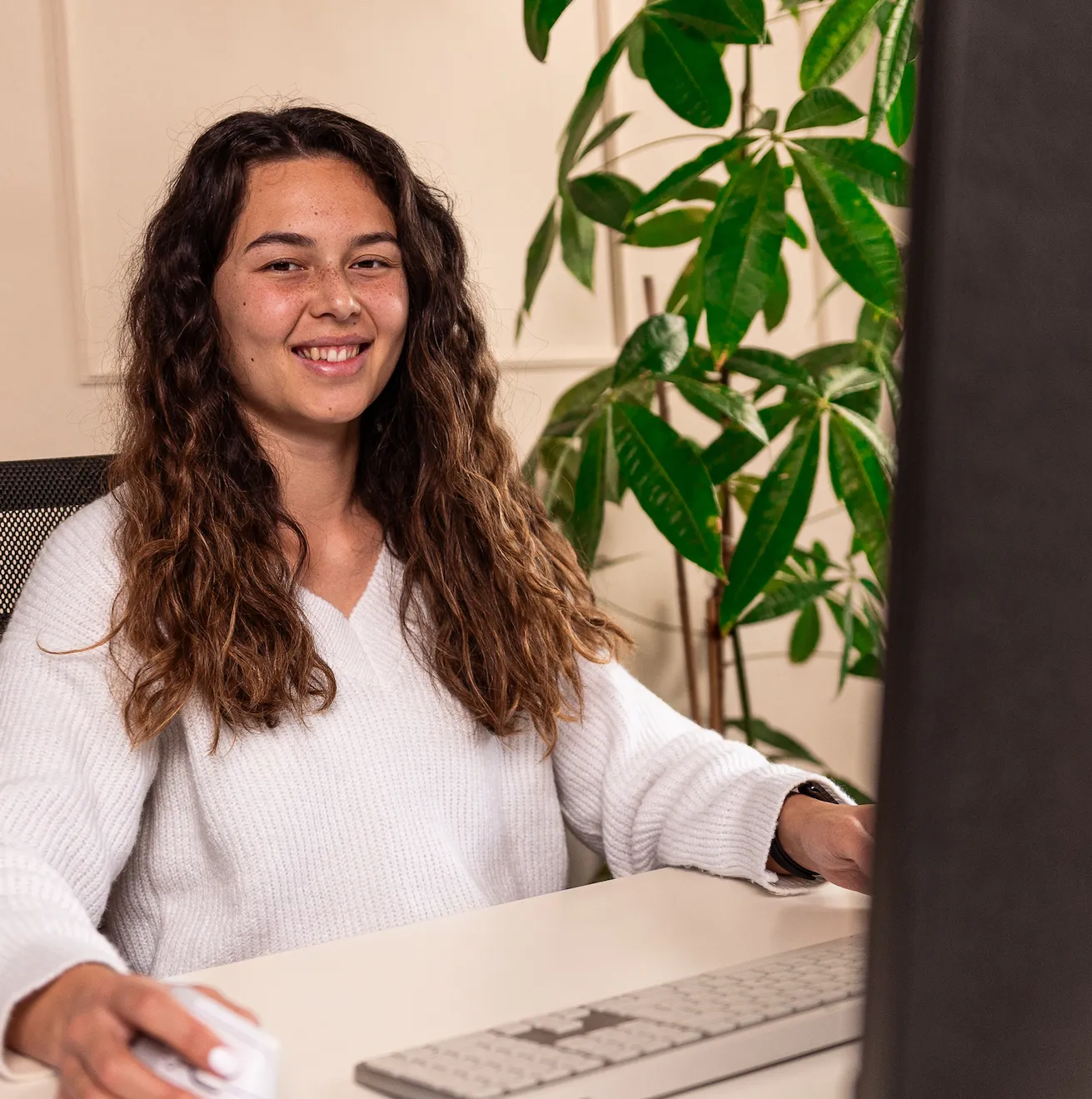Die Geschäftsführerin Lorine Sumono mit langem lockigem Haar sitzt lächelnd an einem Schreibtisch vor einem Computer, umgeben von grünen Pflanzen.