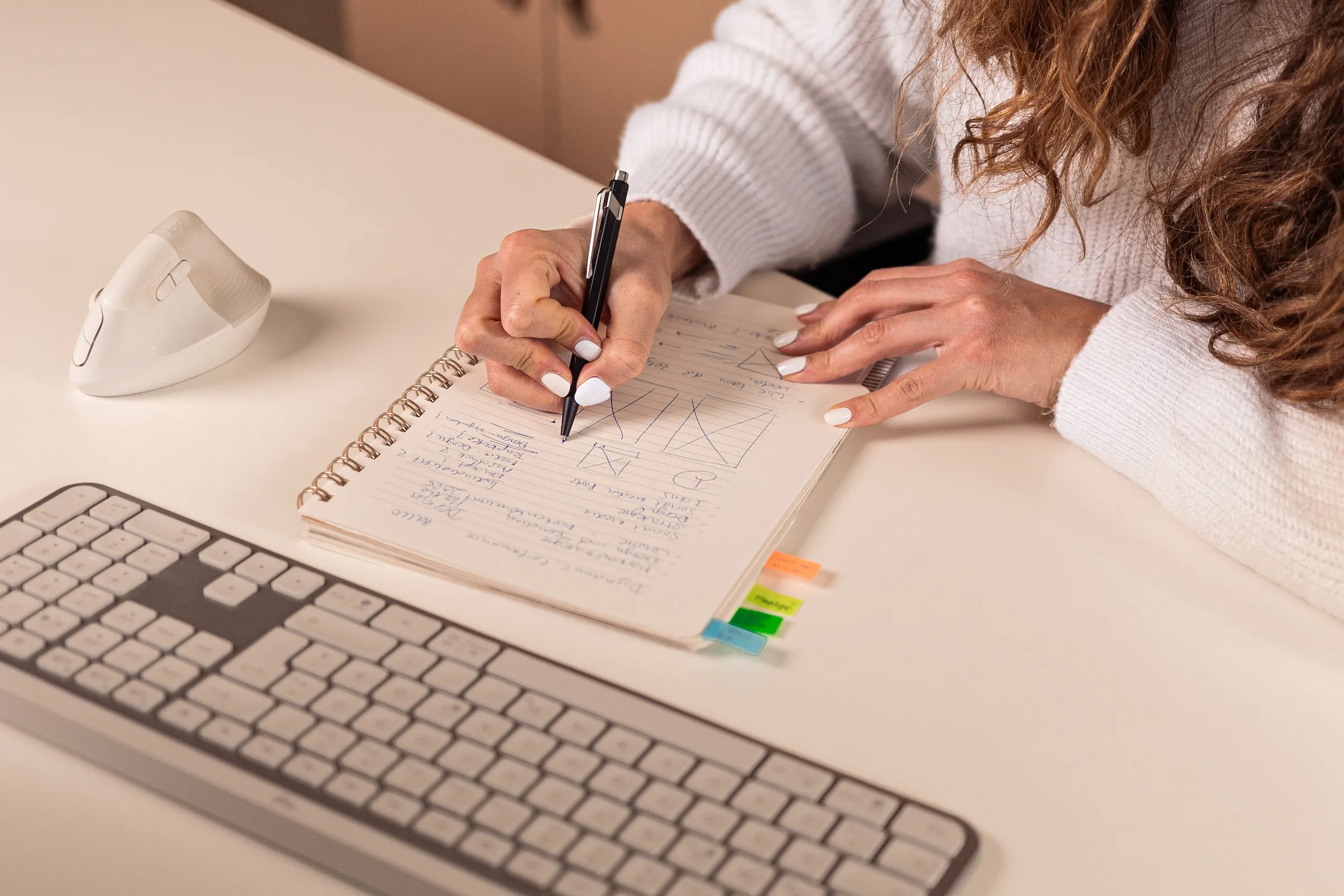 Frau in weißem Pullover schreibt mit einem schwarzen Stift Notizen und Diagramme in ein Spiralnotizbuch auf einem Schreibtisch neben einer Tastatur und einer weißen Maus.