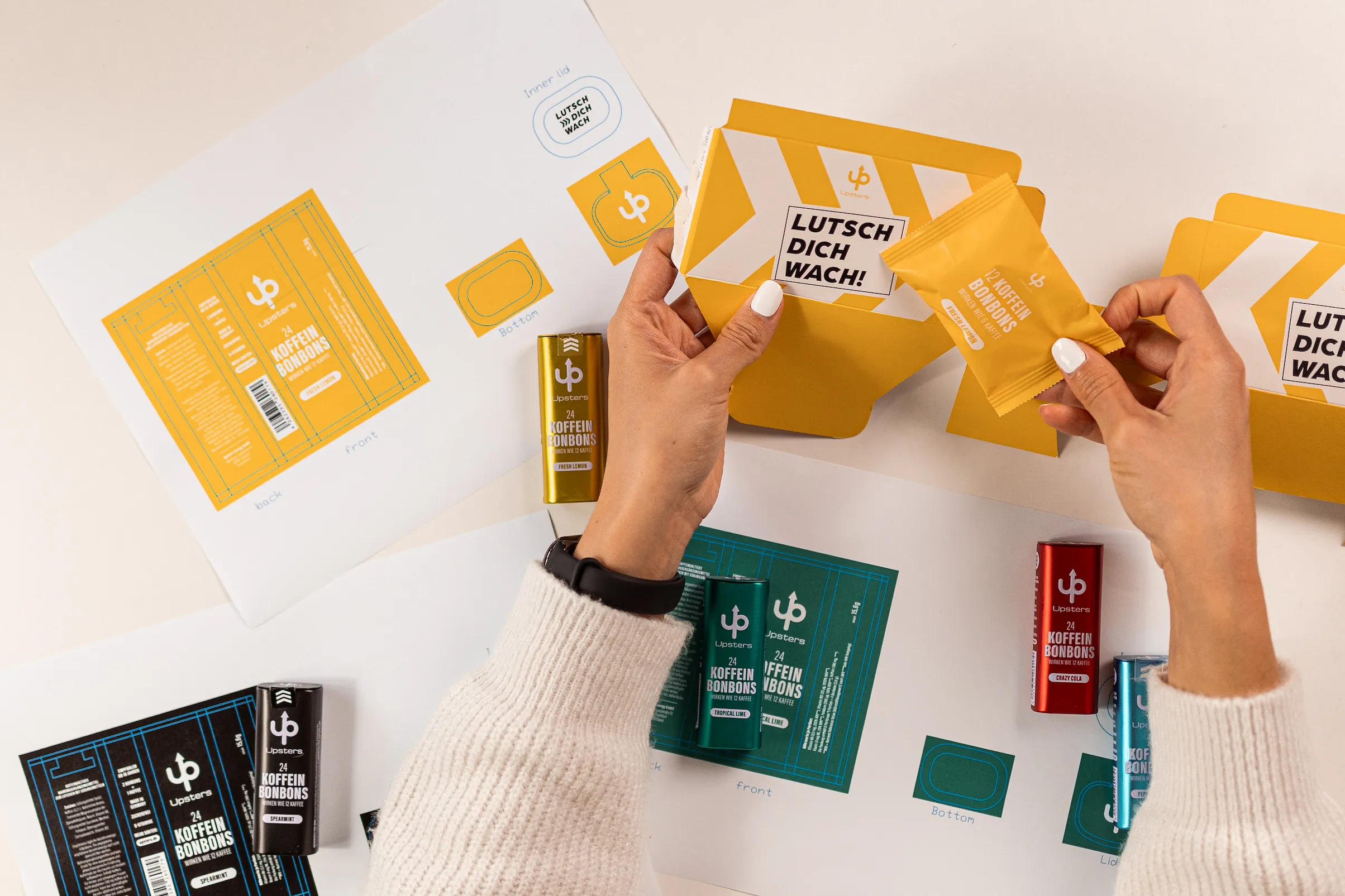 Hände halten eine gelbe Verpackung und Bonbonpackung mit Aufdruck ‚Lutsch dich wach!‘, umgeben von mehrfarbigen Koffeinbonbonverpackungen und Designvorlagen.