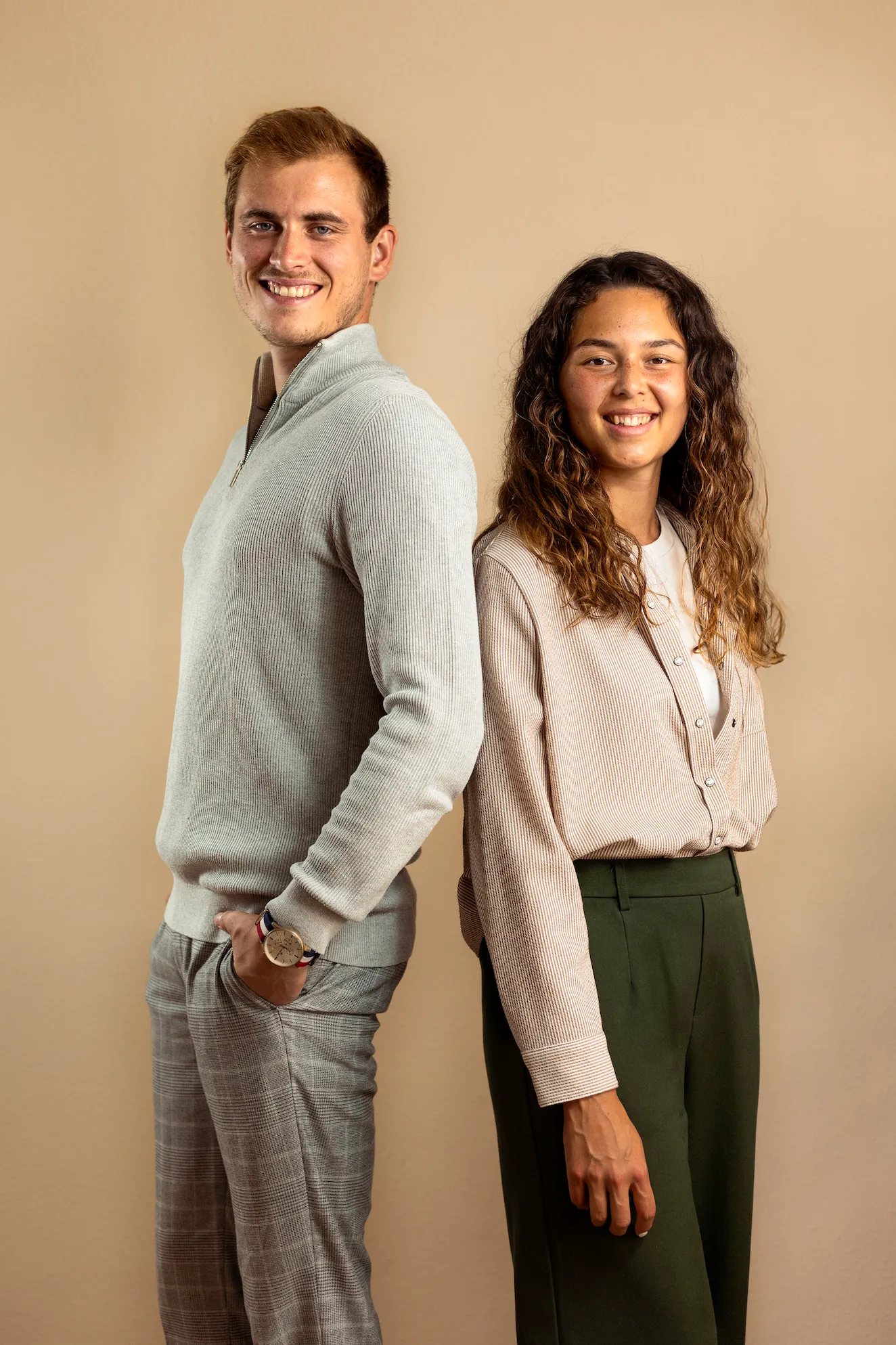 Ein Mann und eine Frau lehnen mit dem Rücken aneinander und lächeln in die Kamera vor einem einfarbigen beigen Hintergrund.