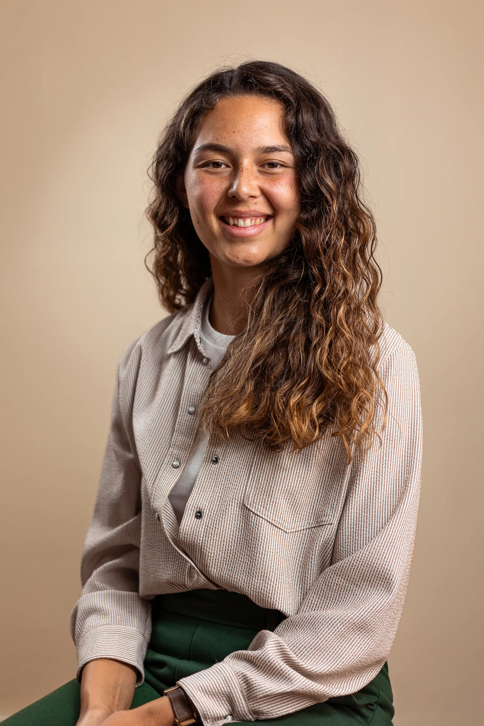 Junge Frau mit langen, lockigen braunen Haaren trägt ein gestreiftes Hemd und lächelt vor beigem Hintergrund.