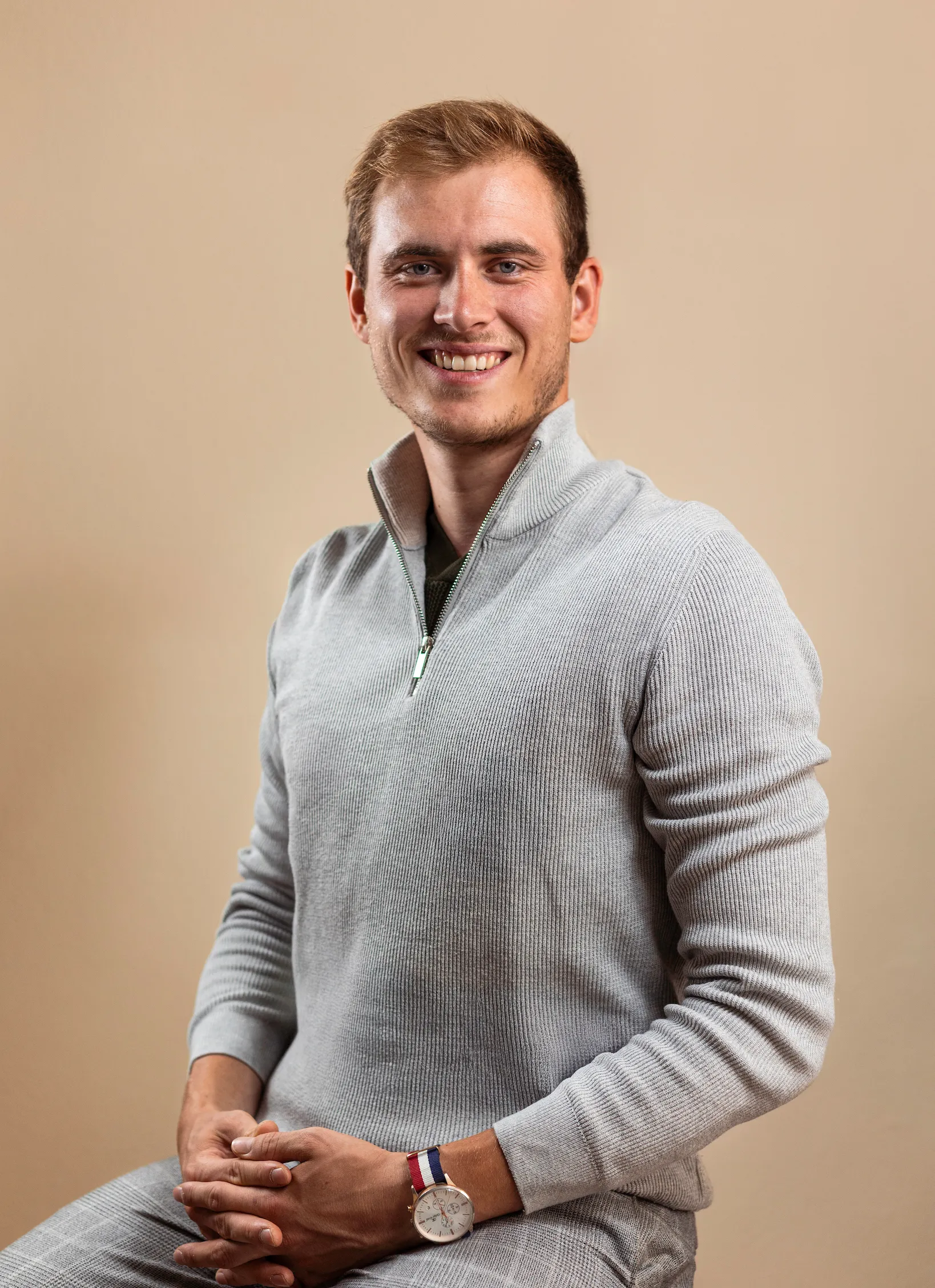 Junger Mann mit grauem Pullover und Armbanduhr sitzt vor hellem Hintergrund und lächelt.