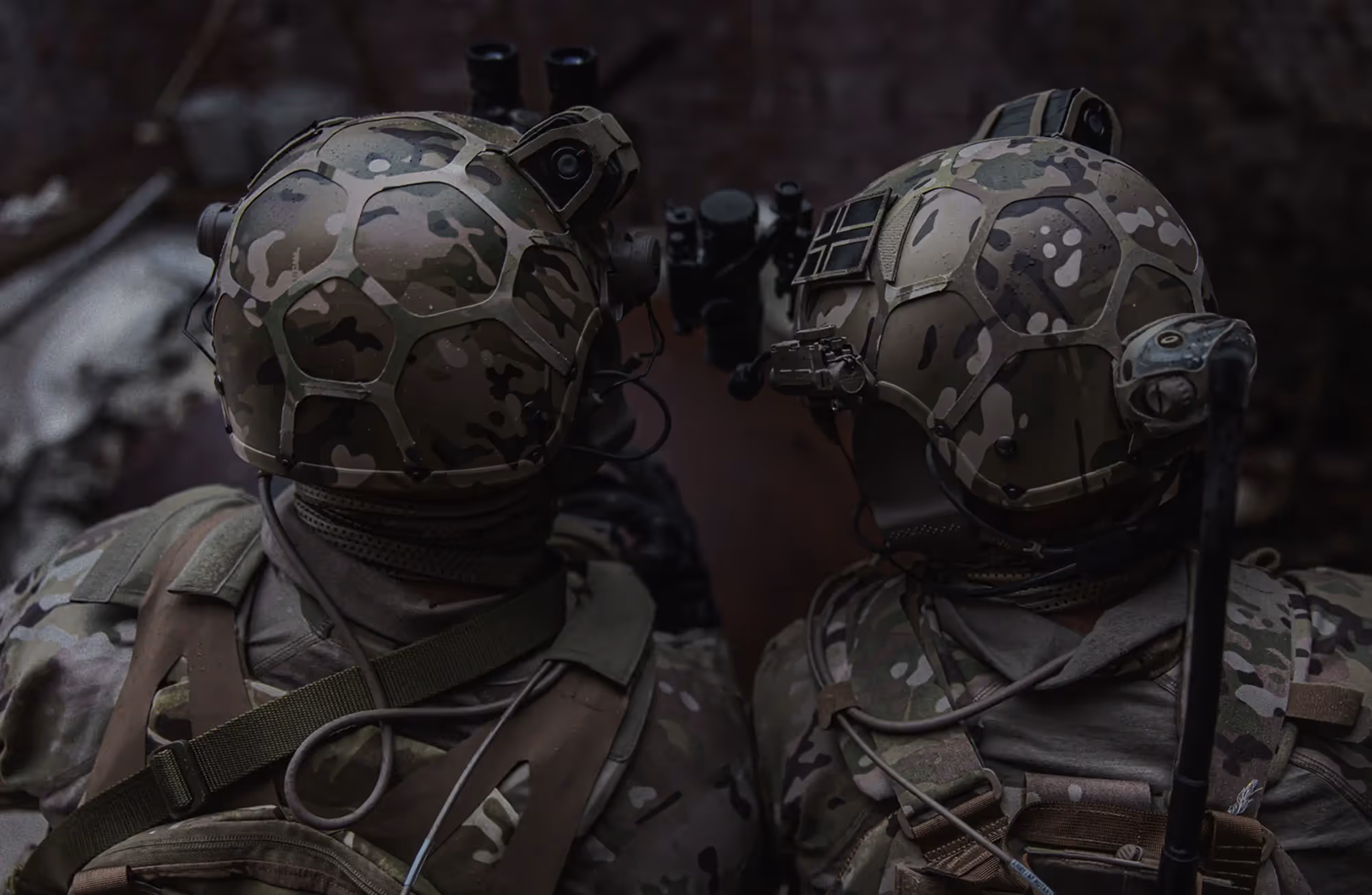 Two soldiers in camouflage gear wearing advanced combat helmets with mounted equipment, viewed from behind in a low-light environment.
