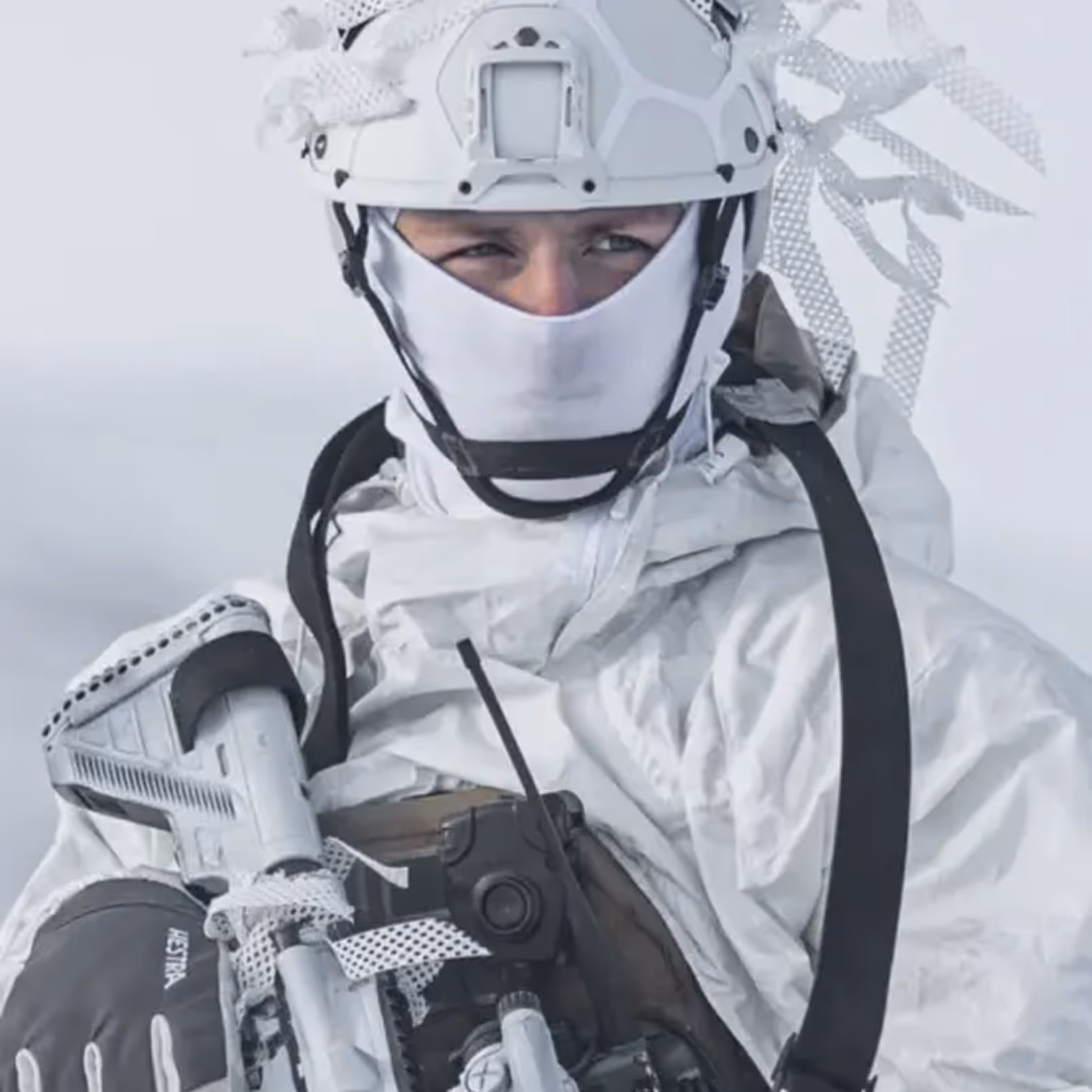 Soldier wearing white winter camouflage gear and helmet, holding a camouflaged firearm in snowy environment.