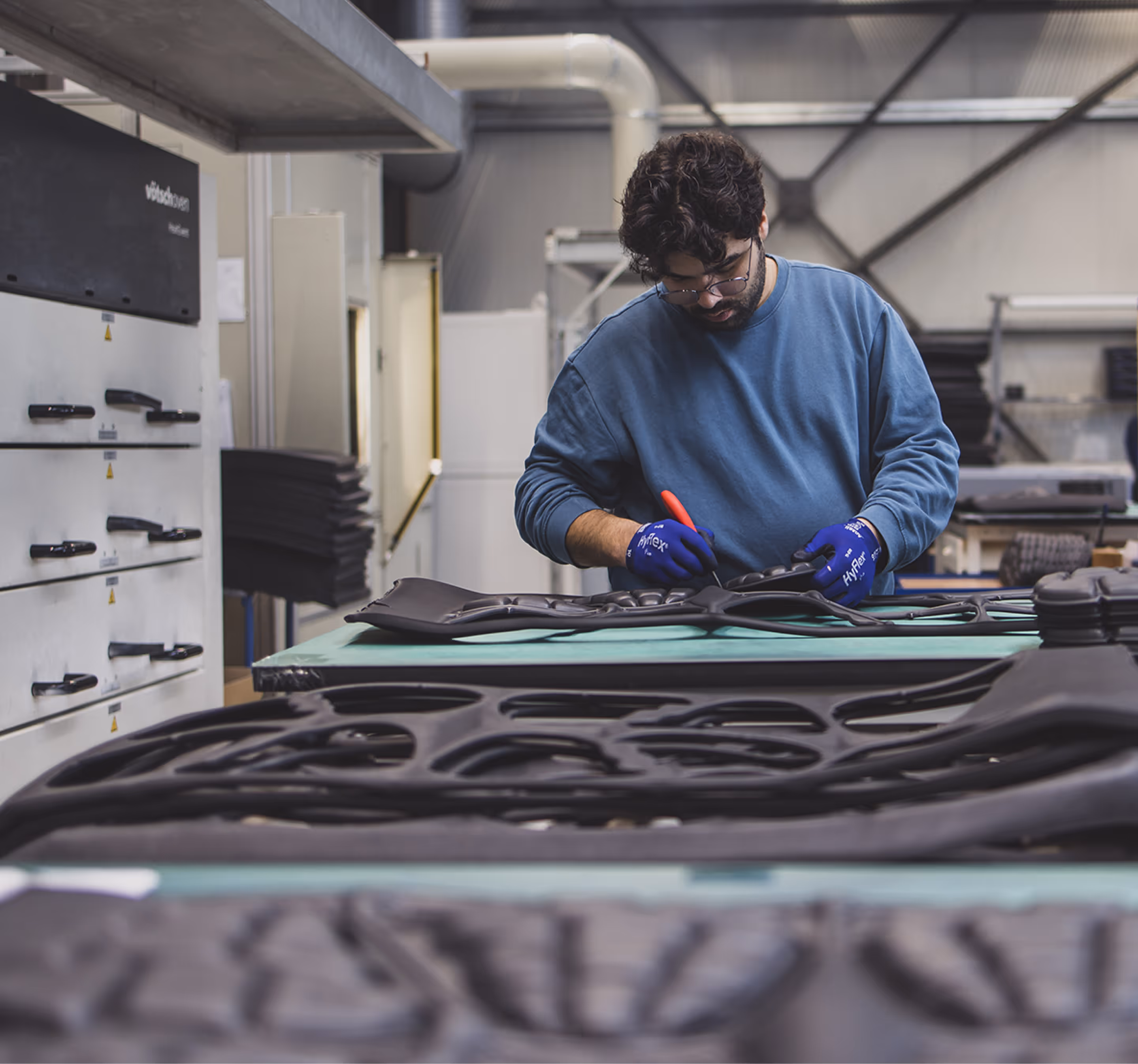 Man wearing gloves and a blue sweatshirt working on black foam or rubber material in a workshop.