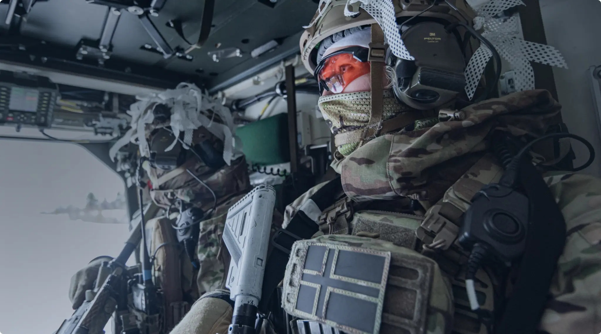 Two soldiers in camouflage gear and helmets sitting inside a military vehicle, one wearing orange-tinted goggles and communicating via headset.