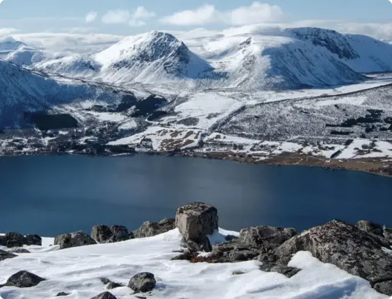 Snow-covered rocky foreground overlooking a lake with snow-capped mountains in the background under a partly cloudy sky.